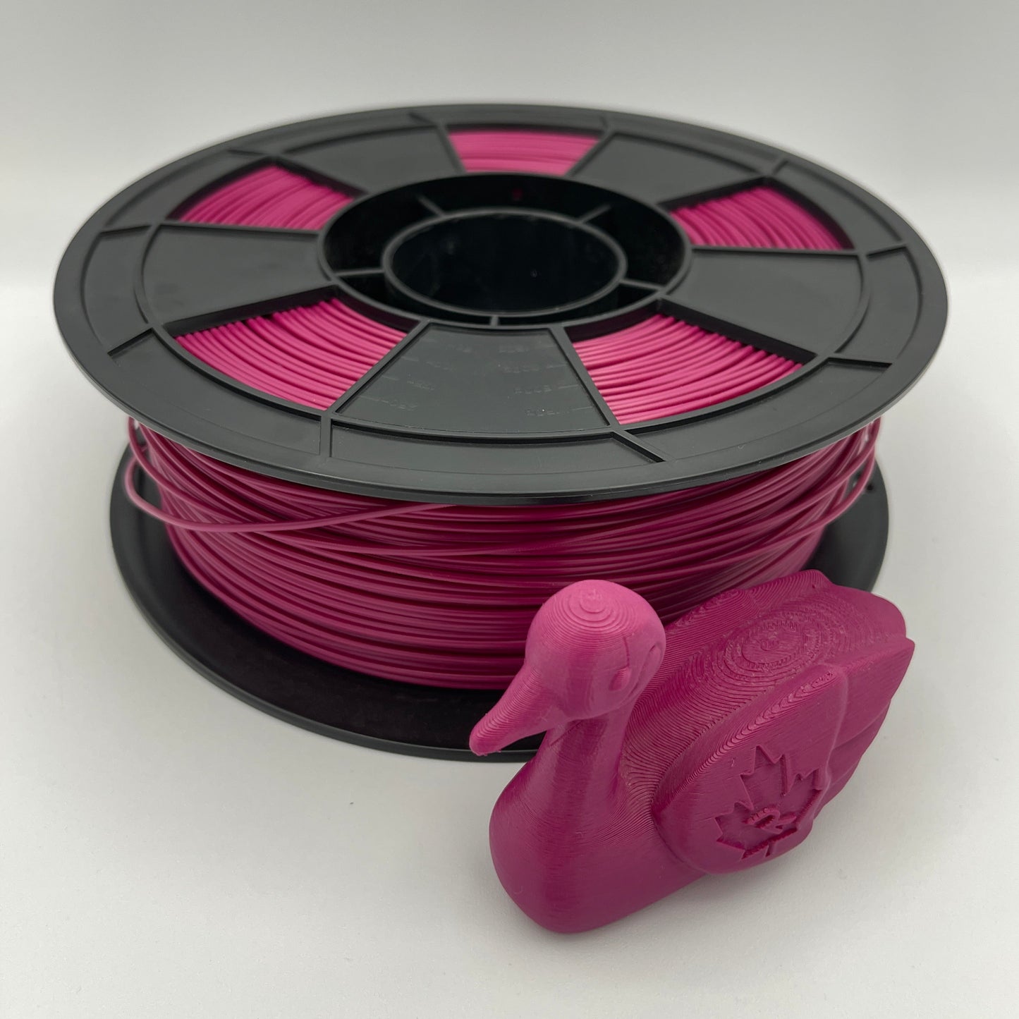 A spool of bright pink 3D printer filament with a black plastic core, shown alongside a small 3D-printed duck model on a clean white background.