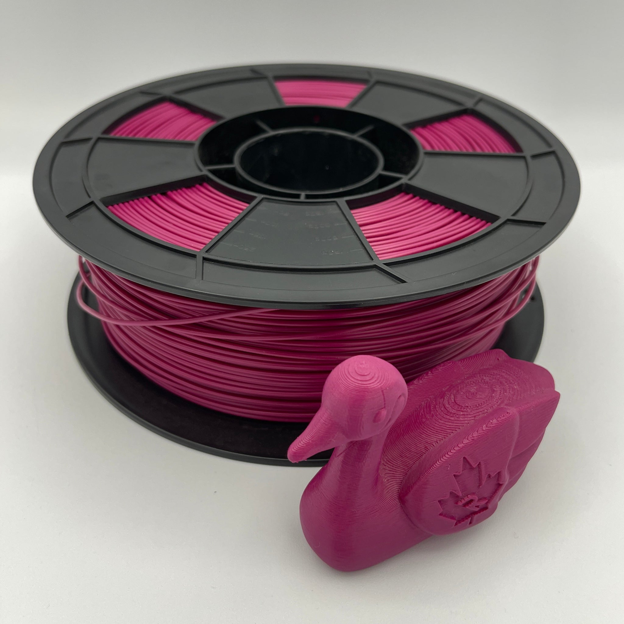 A spool of bright pink 3D printer filament with a black plastic core, shown alongside a small 3D-printed duck model on a clean white background.