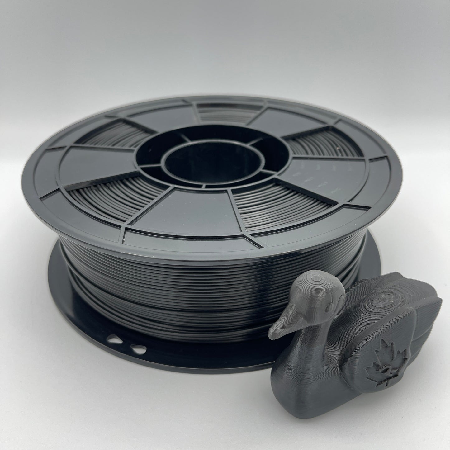 A spool of matte black 3D printer filament with a small duck-shaped sample printed from the filament, displayed on a light gray background.