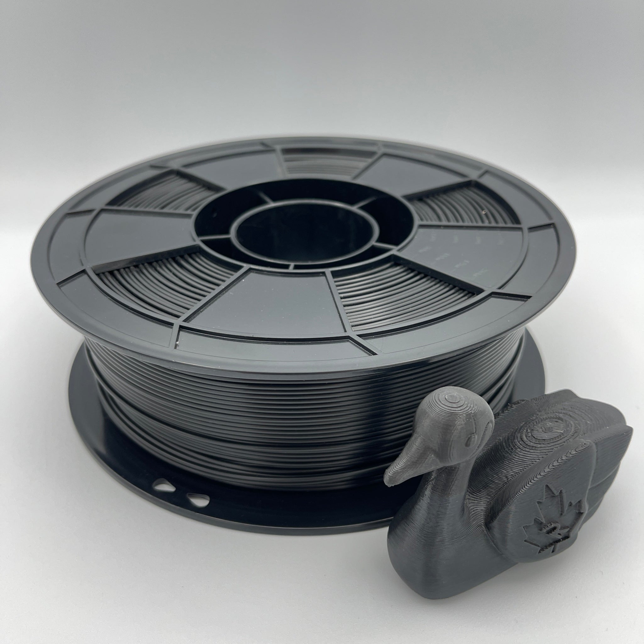 A spool of matte black 3D printer filament with a small duck-shaped sample printed from the filament, displayed on a light gray background.