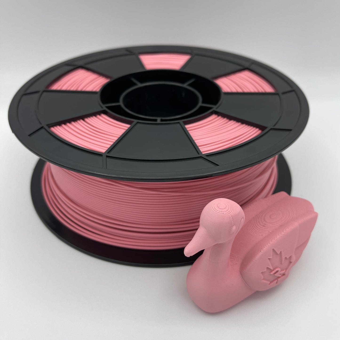 A spool of pink 3D printer filament with a small pink duck-shaped object placed in front, photographed on a clean light gray studio background.