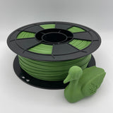 A spool of green 3D printing filament with a small green duck-shaped object placed in front, photographed on a clean light gray studio background.