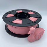 A spool of 3D printing filament in a dusty pink color, wound on a black plastic spool, with a small 3D-printed model of a frog used as a demonstration prop, set against a plain light gray background.