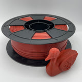 A spool of red 3D printing filament with a small red duck-shaped object placed in front, photographed on a clean light gray to white studio background.