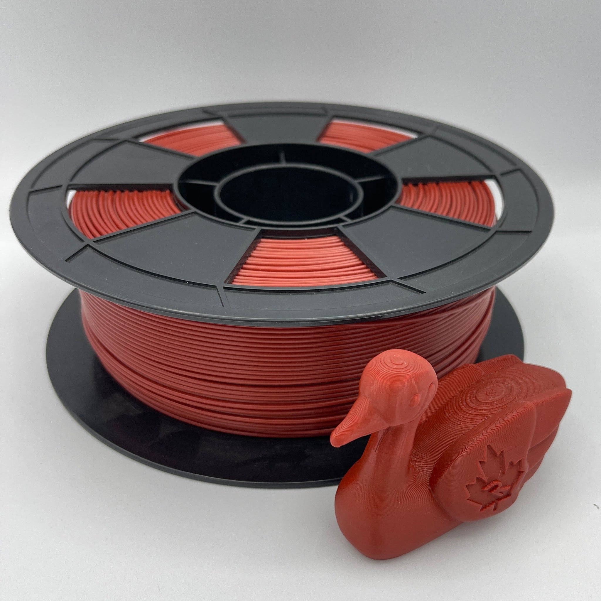 A spool of red 3D printing filament with a small red duck-shaped object placed in front, photographed on a clean light gray to white studio background.