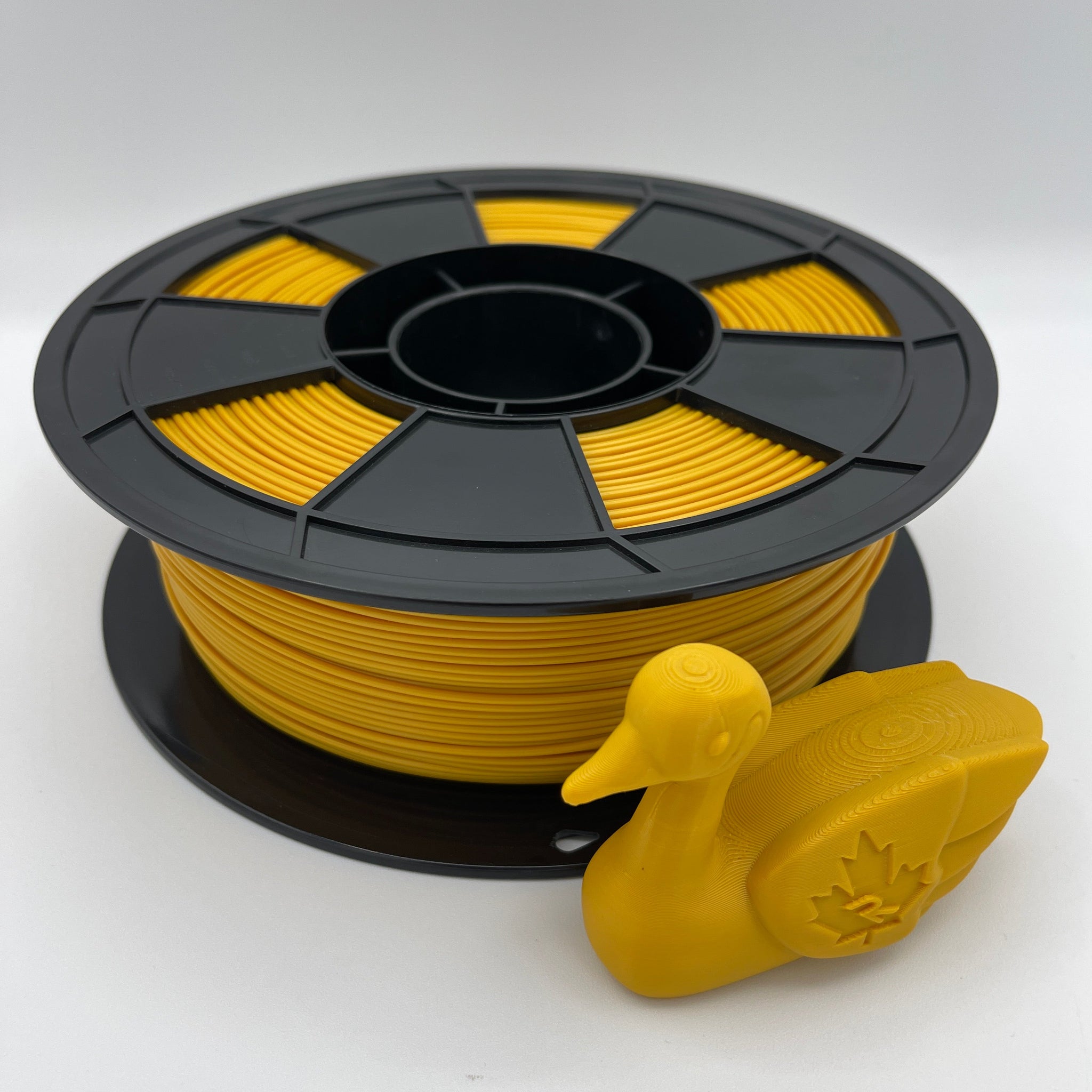 A spool of bright yellow 3D printer filament with a small yellow duck-shaped object placed in front, photographed on a clean light gray studio background.