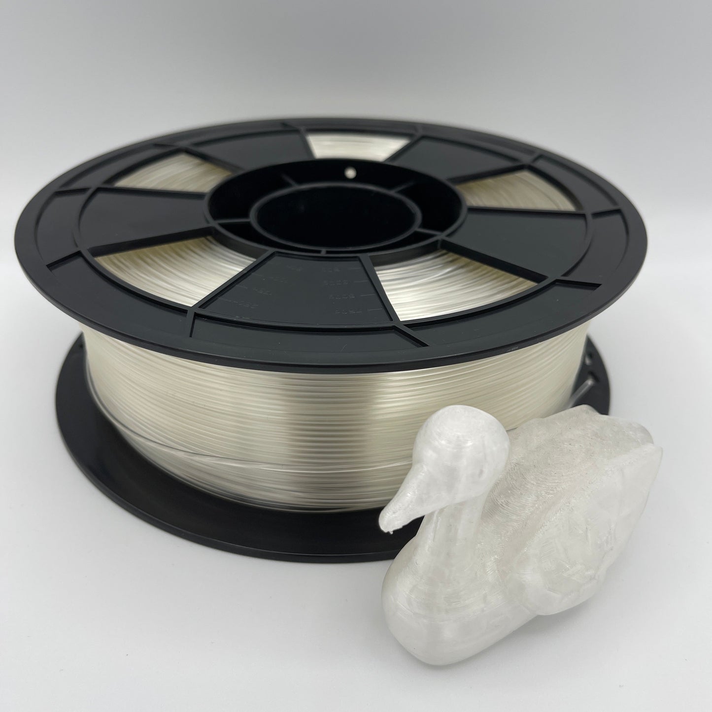 A spool of clear 3D printing filament wound on a black plastic reel, shown next to a small white 3D-printed duck figurine on a light gray background.