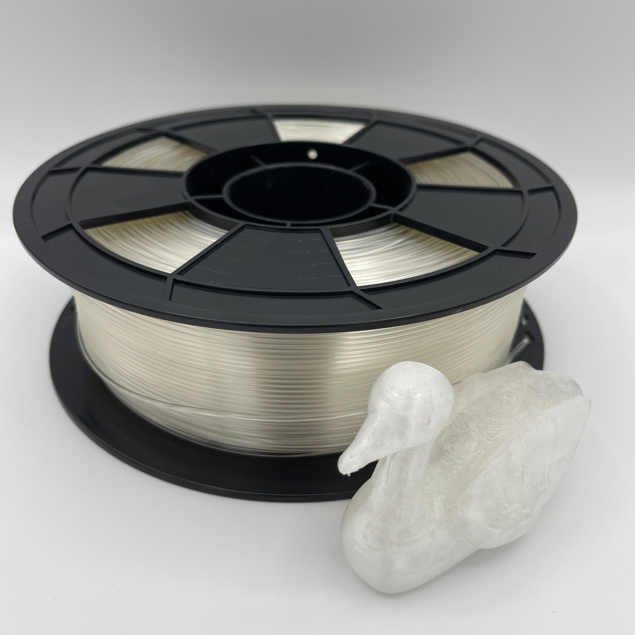 A spool of clear 3D printing filament wound on a black plastic reel, shown next to a small white 3D-printed duck figurine on a light gray background.