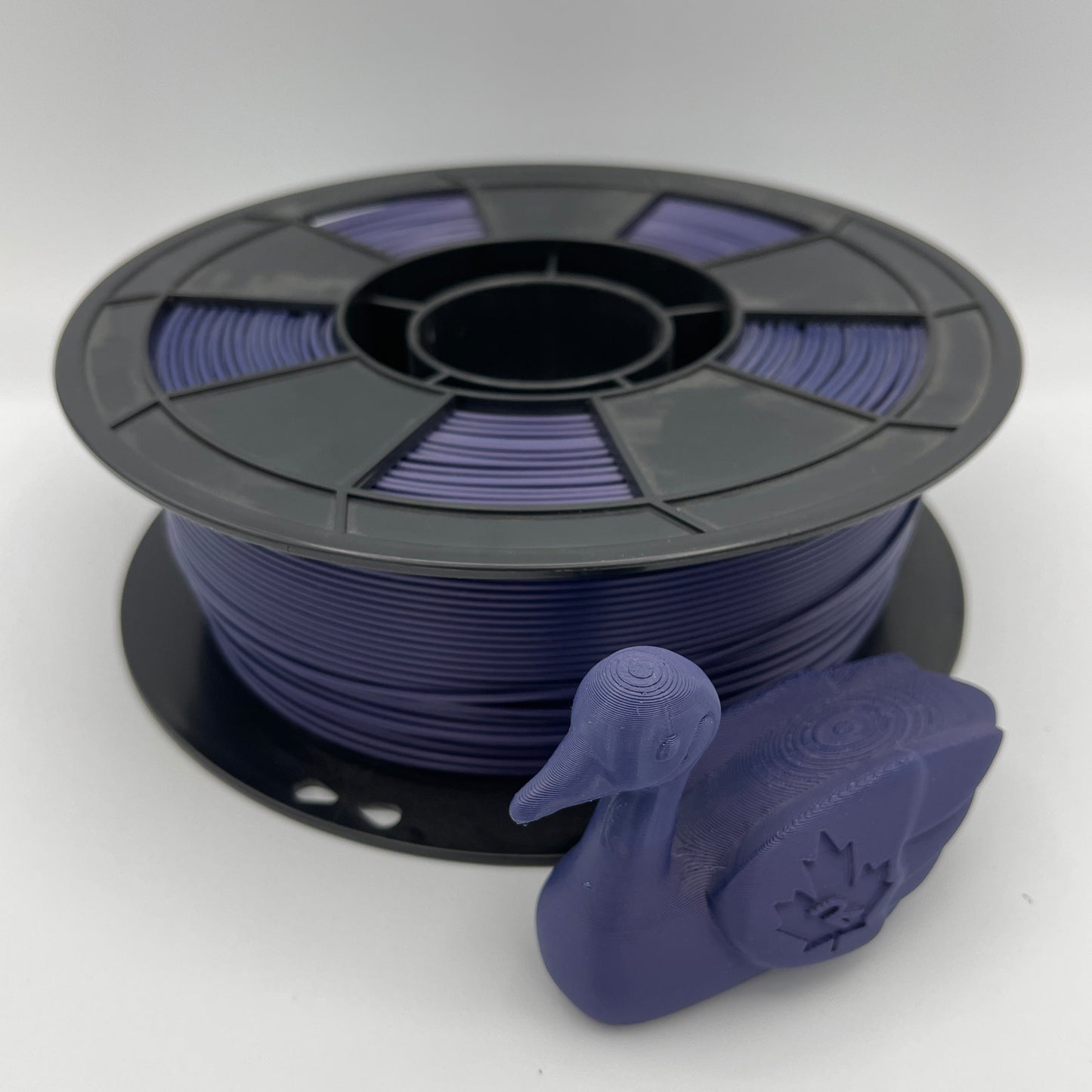 A spool of 3D printer filament wound with dark purple/plum filament, shown next to a small 3D-printed duck model on a light gray studio background.