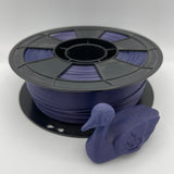 A spool of 3D printer filament wound with dark purple/plum filament, shown next to a small 3D-printed duck model on a light gray studio background.