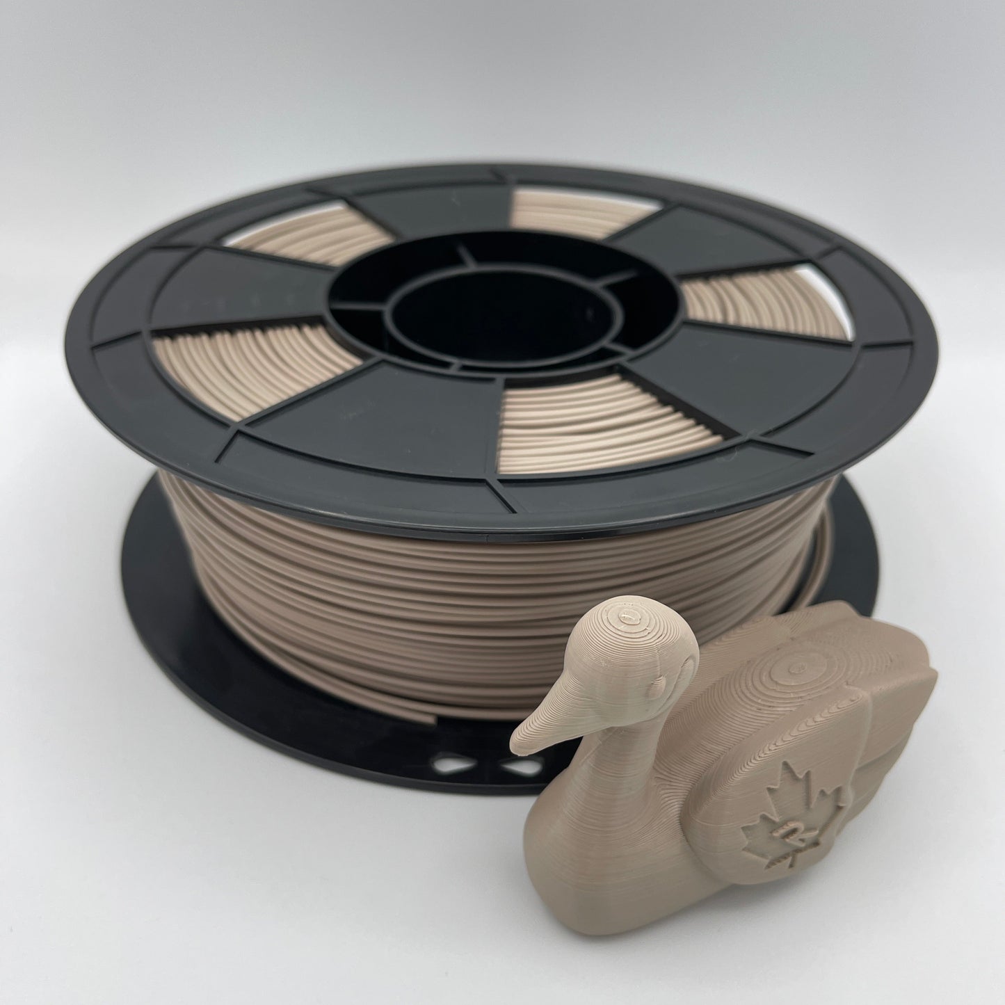A spool of 3D printing filament in a warm beige/tan color, shown next to a small molded duck-shaped object on a light gray studio background.