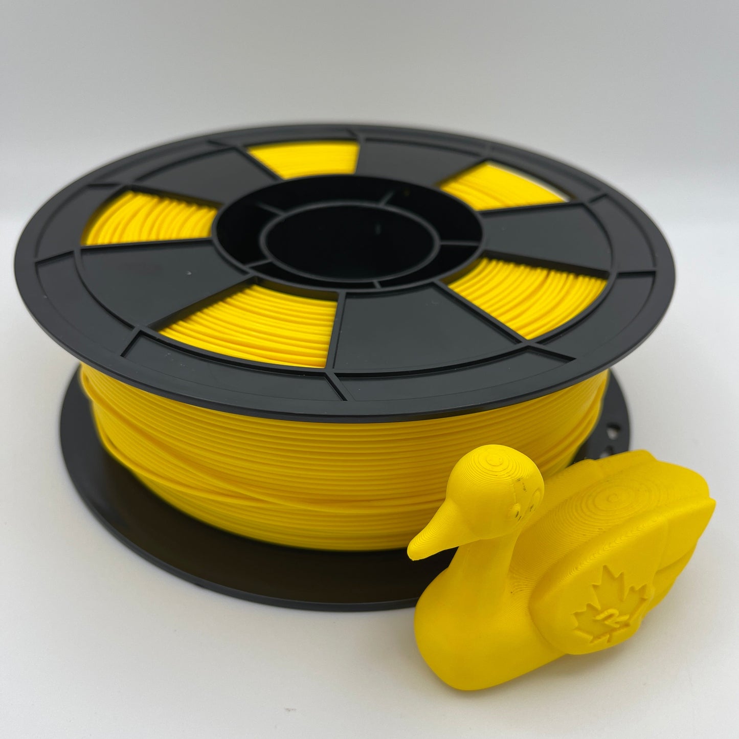 A spool of bright yellow 3D printing filament with a small yellow duck-shaped object placed in front, photographed on a clean white studio background.