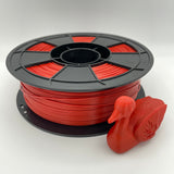 A spool of bright red 3D printing filament with a small red duck-shaped object placed in front, photographed on a clean light gray studio background.