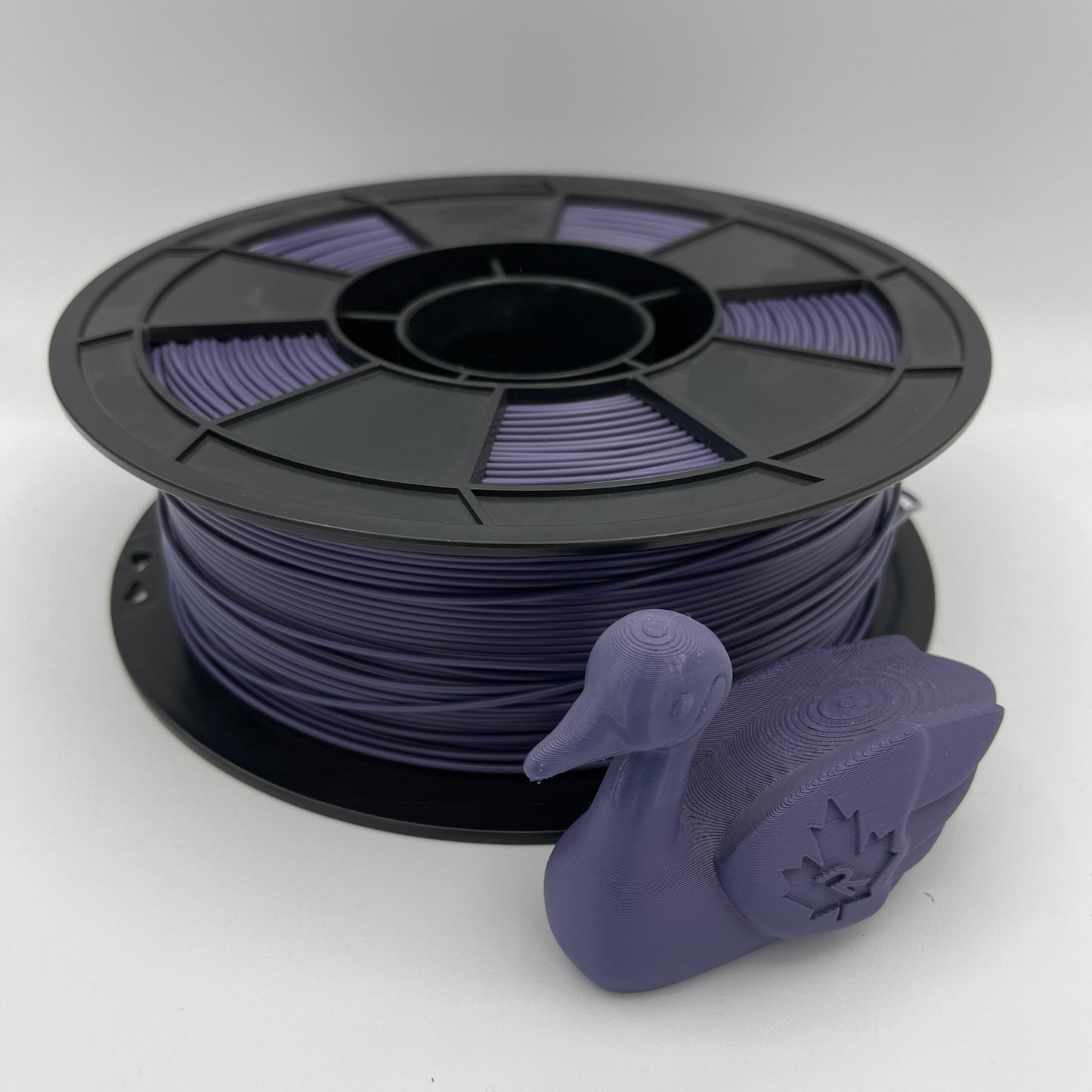 A spool of purple 3D printer filament with a small purple duck-shaped object placed in front, photographed on a clean light gray studio background.