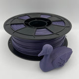 A spool of purple 3D printer filament with a small purple duck-shaped object placed in front, photographed on a clean light gray studio background.