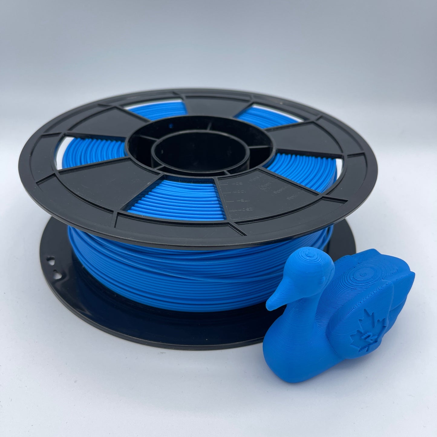 A spool of bright blue 3D printing filament wound on a black plastic core, shown against a clean light gray background with a small blue 3D print sample protruding from the top right.