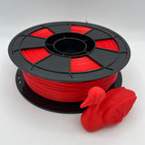 A spool of bright red 3D printer filament with a black plastic core, shown next to a small red duck-shaped 3D print on a light gray background.