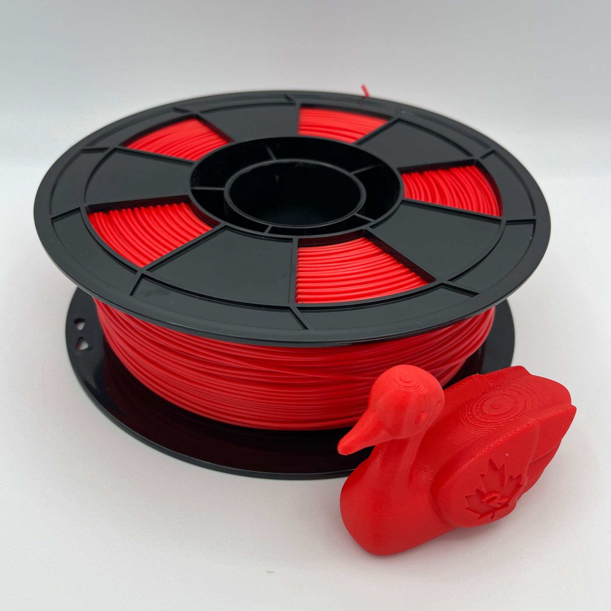 A spool of bright red 3D printer filament with a black plastic core, shown next to a small red duck-shaped 3D print on a light gray background.