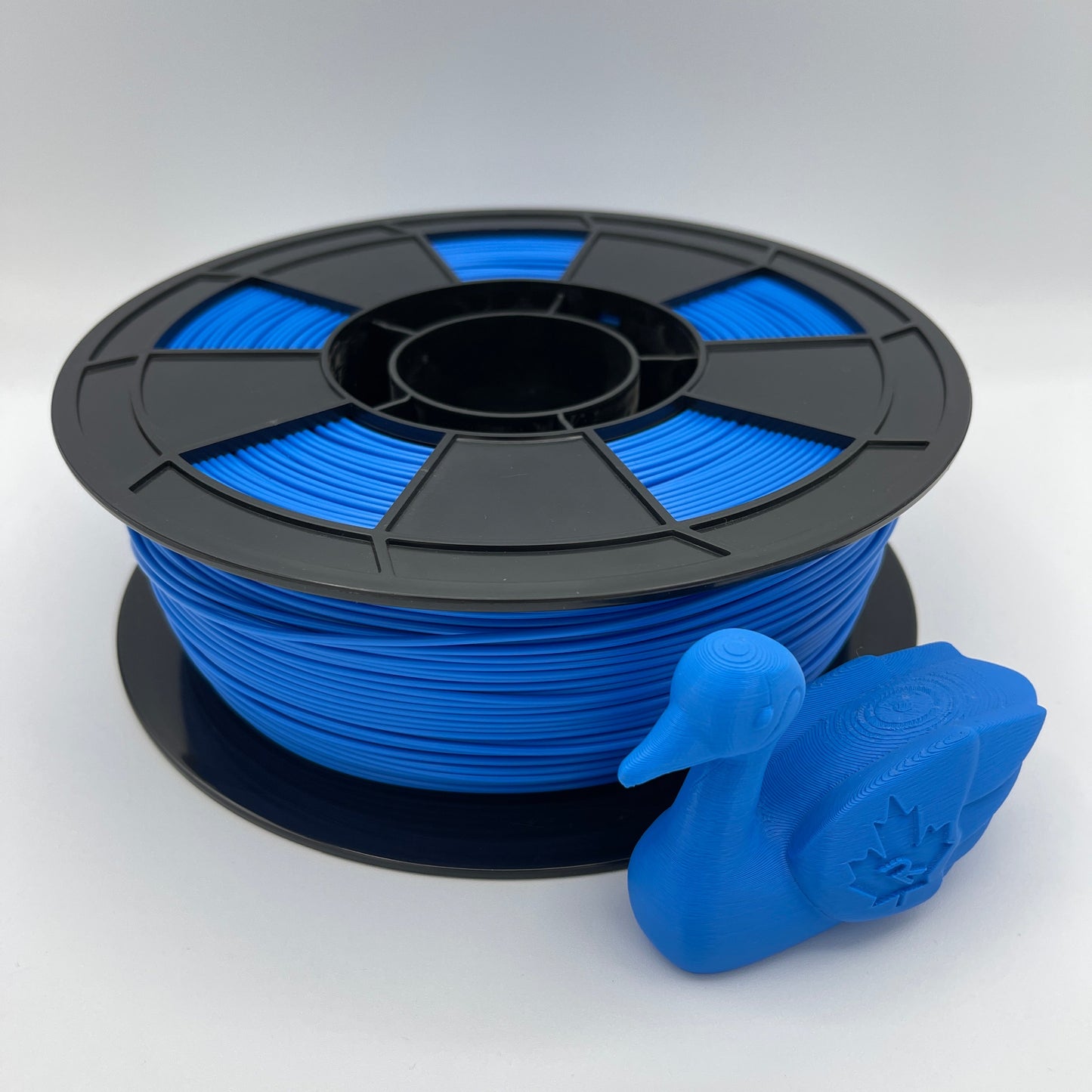 A spool of bright blue 3D printing filament with a black plastic hub, shown next to a small blue duck-shaped printed object on a light gray background.