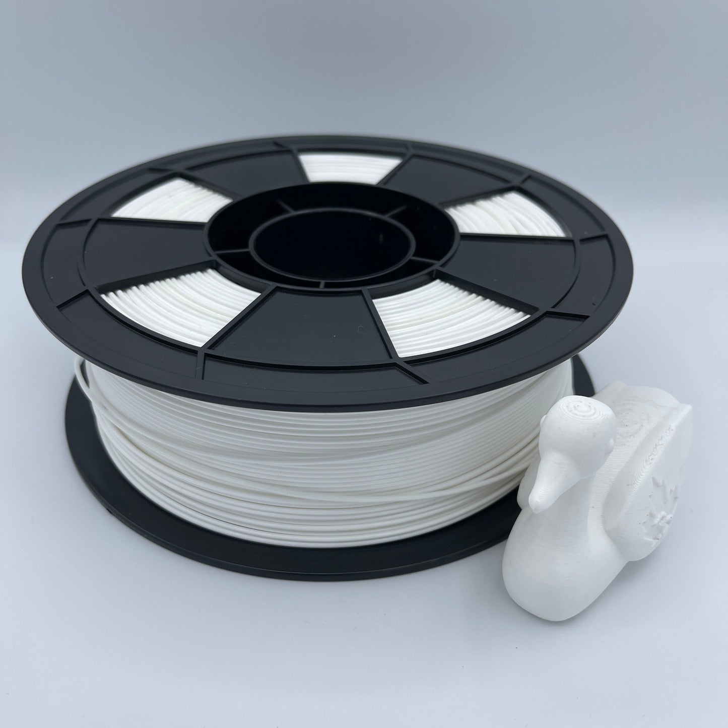 A spool of white 3D printer filament with a black plastic core, shown next to a small white printed duck figurine on a light gray background.