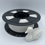 A spool of white 3D printer filament with a black plastic core, shown alongside a small white duck figurine on a light gray studio background.
