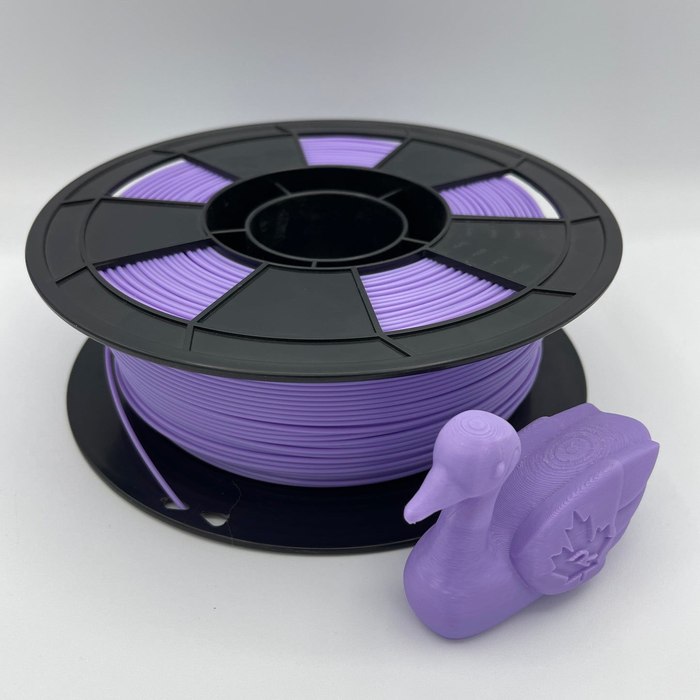 A spool of purple 3D printing filament with a small purple duck-shaped object placed in front, photographed on a clean light gray studio background.