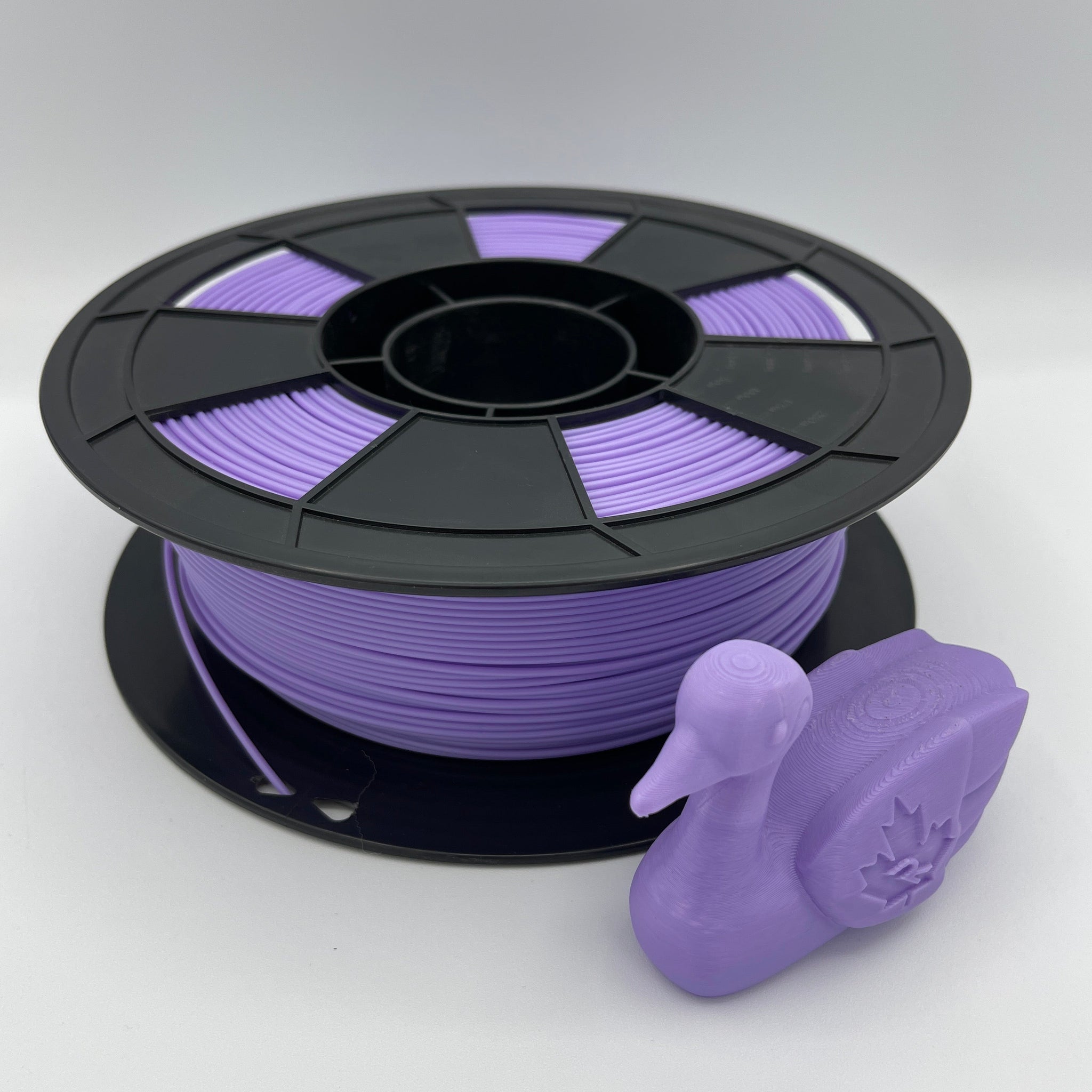 A spool of purple 3D printing filament with a small purple duck-shaped object placed in front, photographed on a clean light gray studio background.