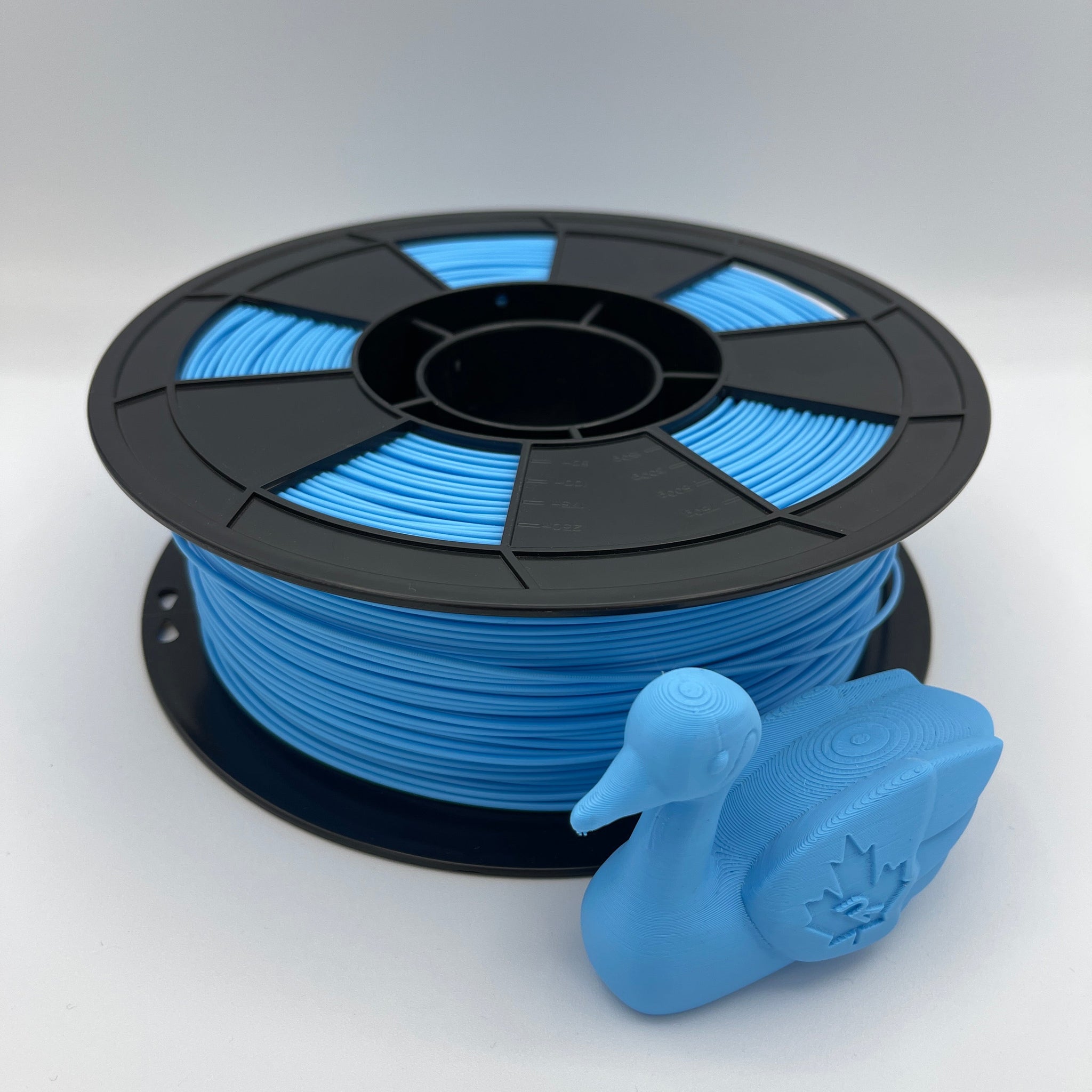 A spool of bright blue 3D printer filament with a black plastic core, shown alongside a small blue duck-shaped printed object on a light gray background.