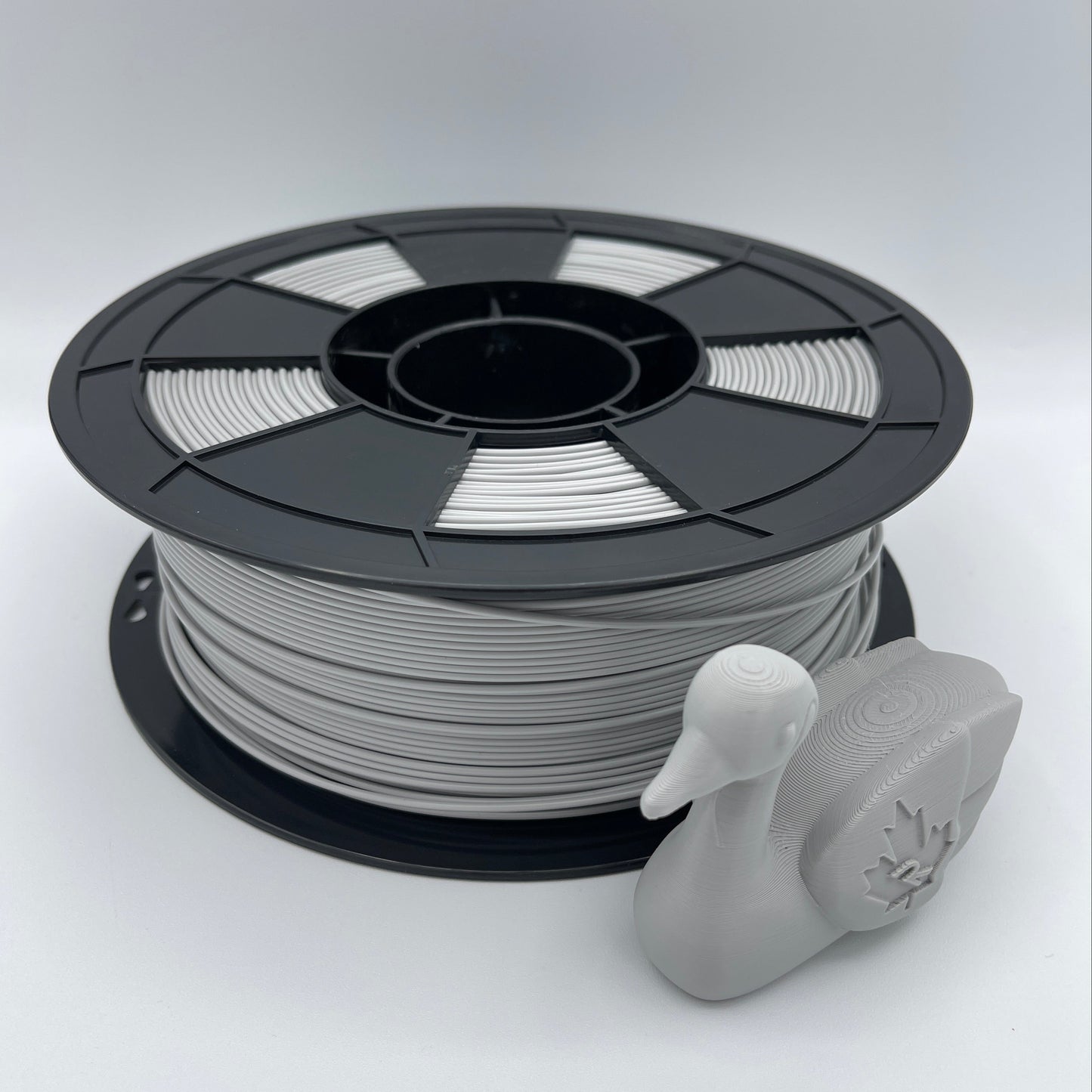 A spool of light gray 3D printing filament with a small gray duck-shaped object placed in front, photographed on a clean light gray studio background.