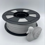 A spool of light gray 3D printing filament with a small gray duck-shaped object placed in front, photographed on a clean light gray studio background.