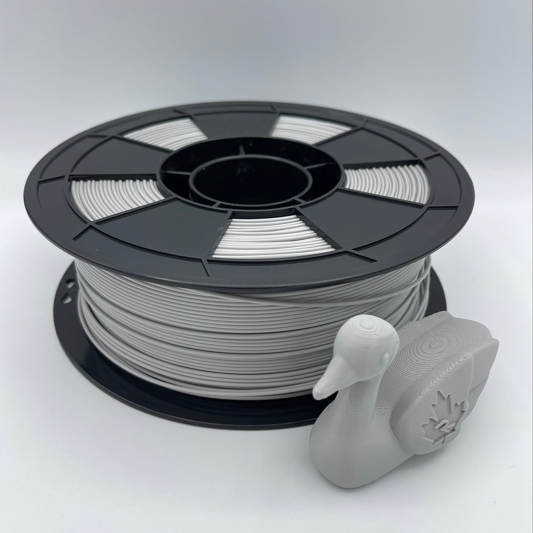 A spool of light gray 3D printing filament with a small gray duck-shaped object placed in front, photographed on a clean light gray studio background.