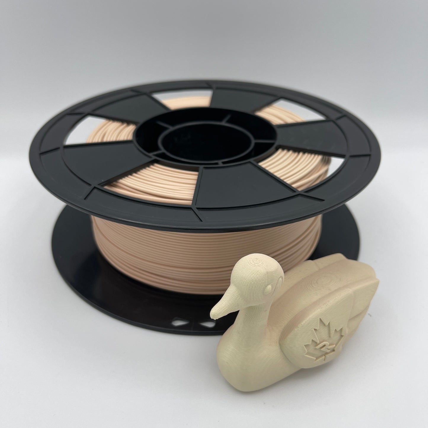A spool of 3D printing filament in a warm beige/tan color, shown next to a small beige duck-shaped object on a clean light gray studio background.