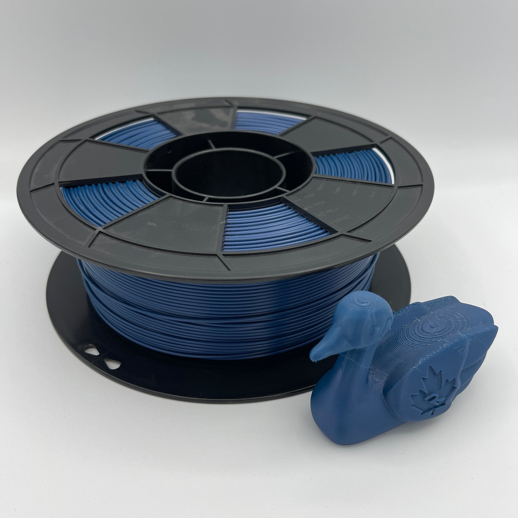 A spool of blue 3D printing filament with a small blue printed duck-shaped object placed in front, photographed on a clean light gray studio background.