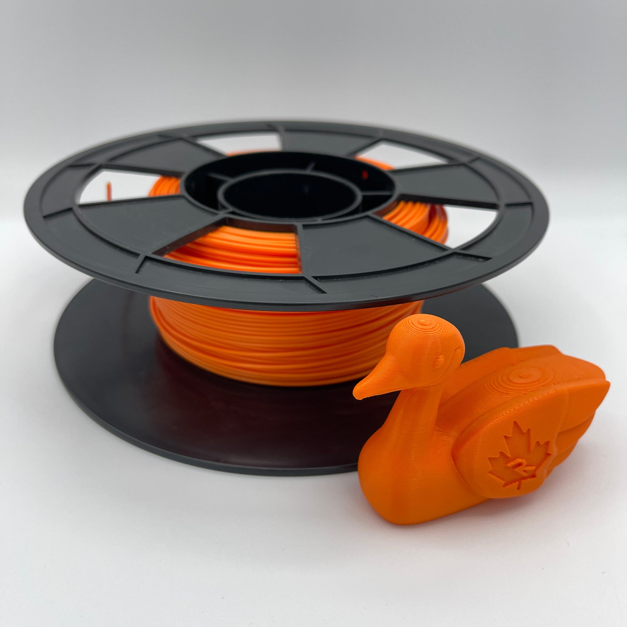 An orange 3D-printed duck figurine placed in front of a black spool wound with matching orange filament, photographed on a clean light gray studio background.