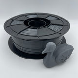 A spool of matte black 3D printer filament with a small gray duck-shaped sample printed on the front, photographed on a clean white studio background.