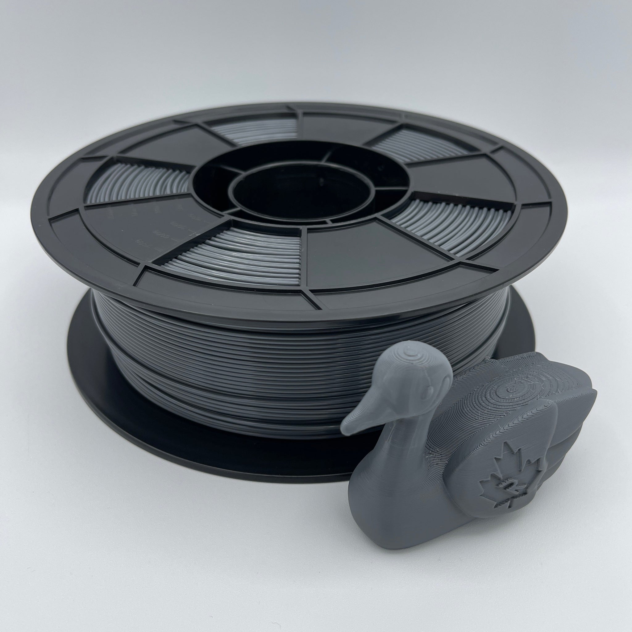 A spool of matte black 3D printer filament with a small gray duck-shaped sample printed on the front, photographed on a clean white studio background.
