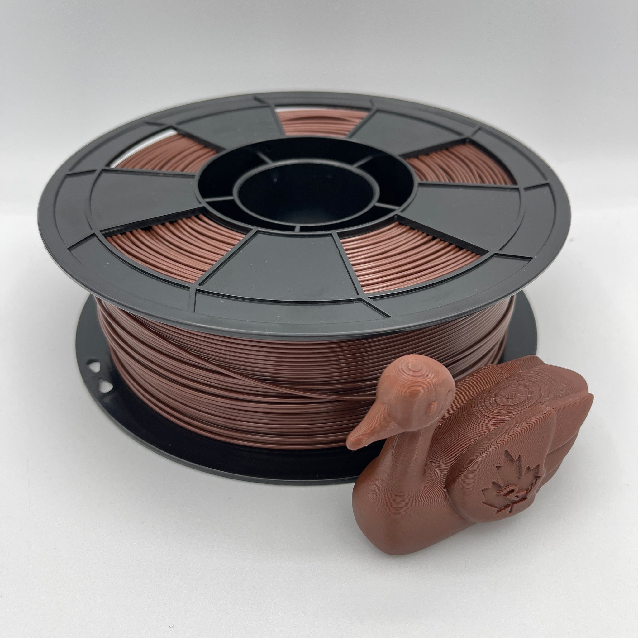A spool of copper-colored 3D printer filament next to a small duck-shaped printed object, photographed on a light gray studio background.